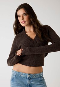 Maglione corto marrone con scollatura a scallop, realizzato in tessuto a maglia testurizzato. Abbinato a jeans blu chiaro con bottoni argentati.