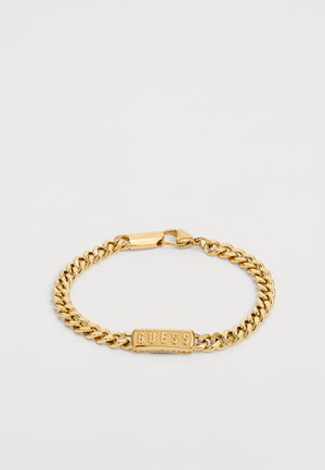 Le bracelet chaîne en or comprend des maillons en curb entrelacés et un charm rectangulaire avec le logo « GUESS » en relief. Détail de fermoir sécurisé visible.