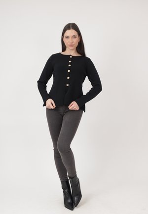 Vrouw staat zelfverzekerd en draagt een zwart vest met knopen, donkergrijze skinny jeans en zwarte puntige enkellaarsjes tegen een effen achtergrond.