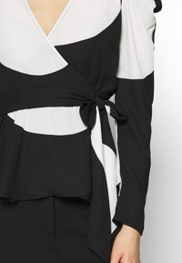Blouse enveloppante noir et blanc à manches longues, avec un lien à la taille, des formes géométriques audacieuses et un décolleté en V profond.
