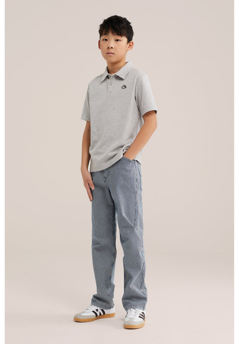 Adolescent debout, une main dans la poche, portant un polo gris, un pantalon bleu rayé et des baskets blanches avec des rayures noires.