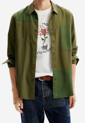 Personne portant une chemise patchwork vert olive ouverte sur un t-shirt blanc avec un motif floral et un jean patchwork bleu.