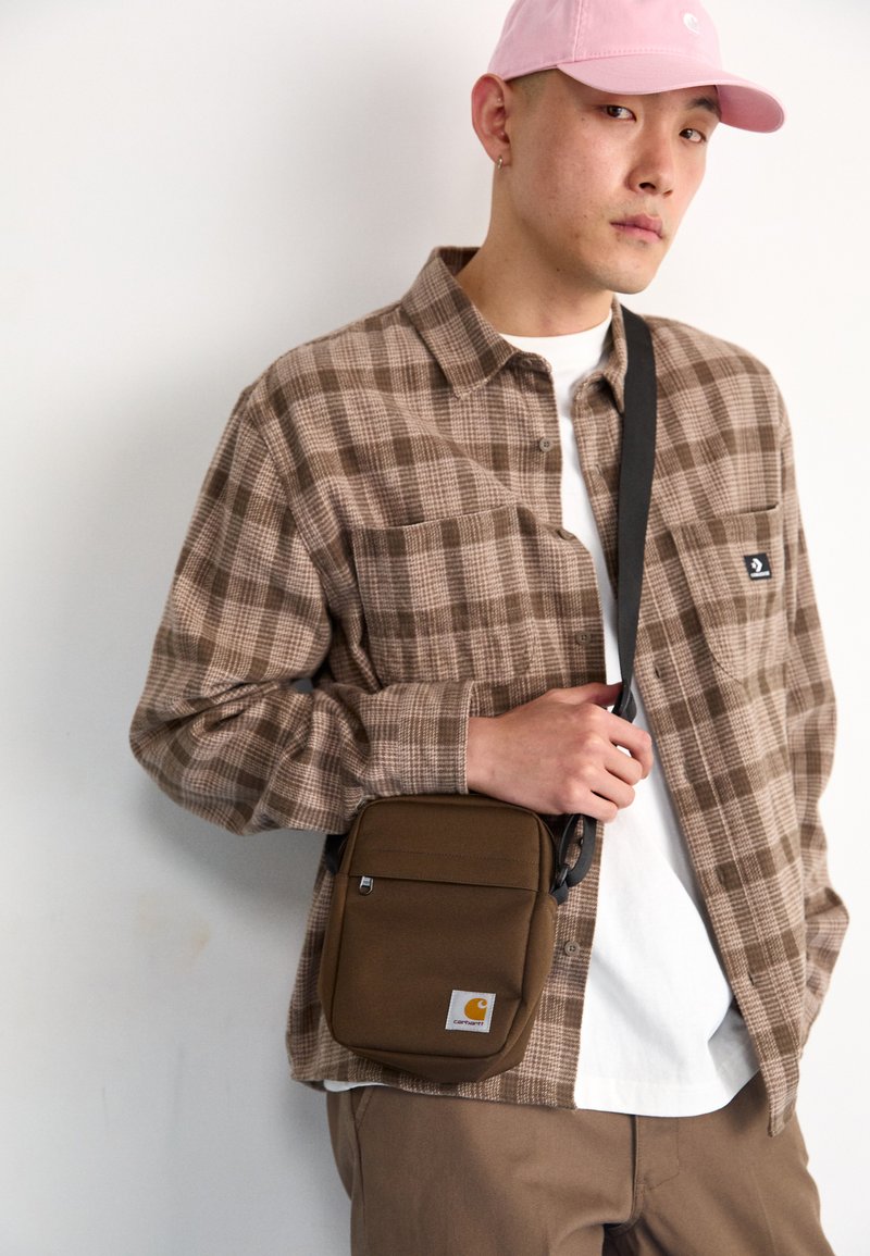 Cămașă cu carouri maro peste un tricou alb, asortată cu pantaloni bej. O geantă crossbody compactă maro cu un logo este ținută la talie. Se poartă o bandă roz.
