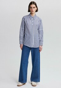 Camisa de botones a rayas en blanco y azul marino con cuello, combinada con pantalones vaqueros de pierna ancha en azul y zapatillas de color claro.