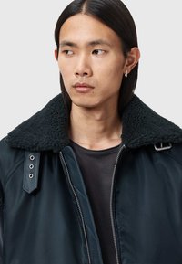 Nuori henkilö, jolla on suorat mustat hiukset ja hopeinen rinkkorengaskoru, pukeutuneena tummaan takkiin, jossa on suuri pörröinen kaulus, yksinkertaista taustaa vasten.