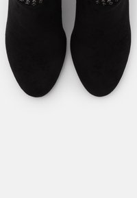 Botas negras hasta el tobillo con una textura de ante suave, punta afilada y un acento decorativo de hardware en el puño. Diseño minimalista.
