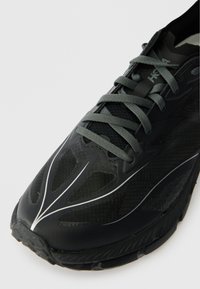 Černá sportovní bota s prodyšnou síťovinou a syntetickým svrškem, vyvýšenými texturovanými vzory, šedými tkaničkami a silnou gumovou podešví. Obsahuje reflexní detaily.