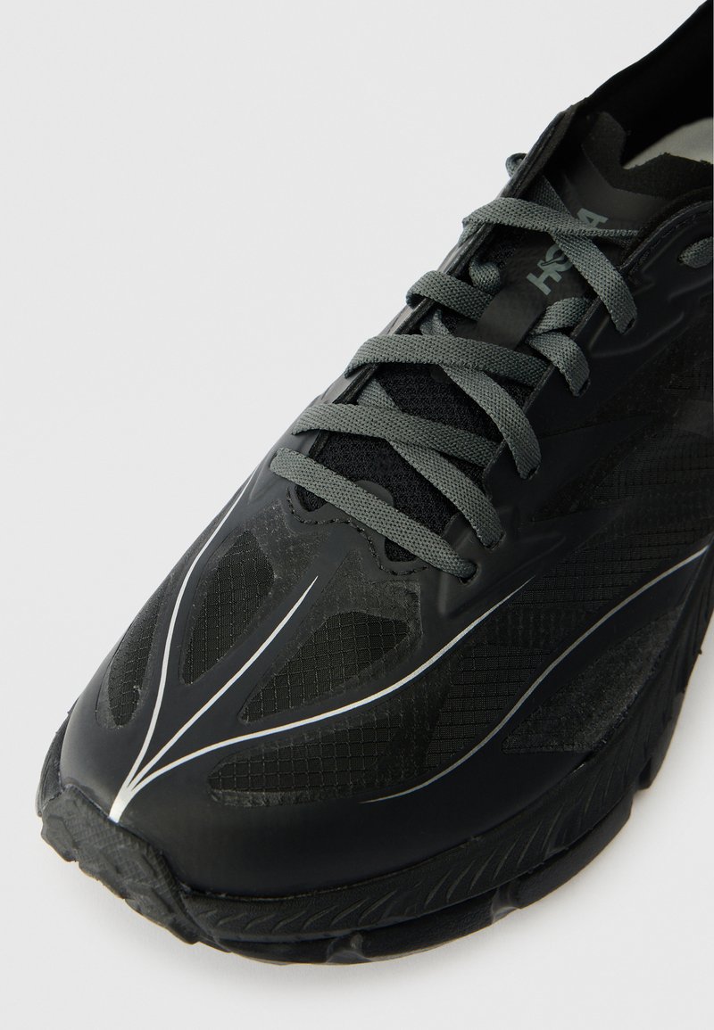 Zwarte sportieve schoen met een bovenwerk van mesh en synthetisch materiaal, verhoogde textuurpatronen, grijze veters en een dikke rubberen buitenzool. Bevat reflecterende accenten.