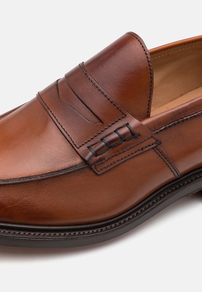 Mocassino in pelle marrone con punta arrotondata, finitura liscia, dettagli cuciti decorativi e un bordo nero lungo la suola.