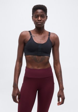 Heas vormis inimene lühikeste juustega, kes kannab musta spordirinnahoidjat ja kõrge vöökohaga bordoopunaseid retuuse, seisab ühetaolise hele taustaga.