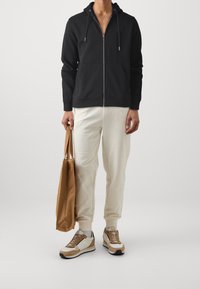 Černá mikina s kapucí a zipem, s šňůrkami, smetanové tepláky a béžové tenisky. Drží hnědou tašku. Minimalistický design a pohodlný střih.