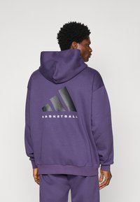Fialová mikina vyrobená z měkké tkaniny, s velkým černým logem Adidas a slovem "BASKETBALL" na zádech. Design s klokaní kapsou.