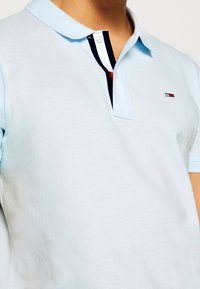 Polo bleu clair avec un col côtelé, doté d'une patte de boutonnage à trois boutons avec des rayures bleu marine, blanches et rouges, et un petit logo sur la poitrine.