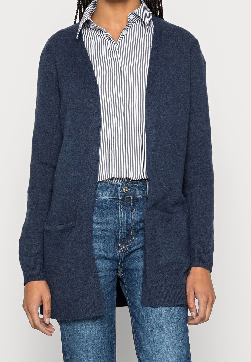 Marineblauer Cardigan, offen vorne mit zwei Fronttaschen, aus weichem Material. Getragen über einem gestreiften Hemd mit Knopfverschluss und Jeans.