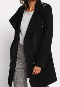 Zwarte jas met een sjaalkraag, textuurstof, voorzakken met rits en een ontspannen pasvorm. Gecombineerd met een grijze trui en geruite broek.