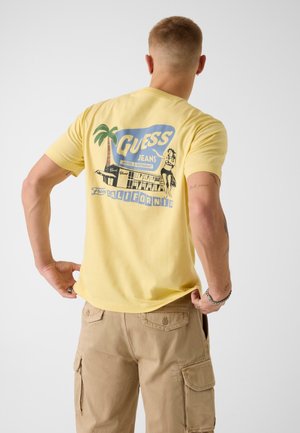 Mężczyzna ubrany w żółtą koszulkę z retro grafiką Guess Jeans California, beżowe krótkie spodnie typu cargo, stojący na jednolitym białym tle.