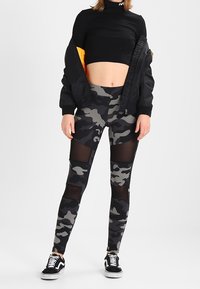 Žena nosí černý cropped rolák, černou bomber jacket s oranžovým podšívkou, kamuflážové legíny s síťovými panely a černé tenisky.