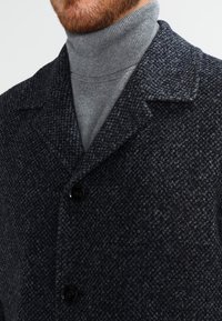 Abrigo de lana texturizada en gris con cuello de pico, que cuenta con tres botones y un sutil patrón de cuadros, llevado sobre un cuello de tortuga gris.