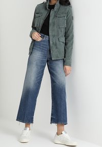 Grijze canvas jas met knoopsluitingen en vier voorzakken, gecombineerd met wijdvallende blauwe jeans en witte sneakers.