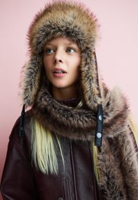 Hnědá kožená pilotní čepice s tlustým kožešinovým lemem a odpovídajícím šálou z umělé kožešiny, s černými knoflíky a měkkými, strukturovanými materiály.