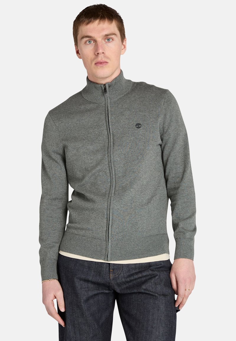 Młody mężczyzna w szarej rozpinanej bluzie z wysokim kołnierzem i ciemnych dżinsach, stojący na tle gładkiej białej ściany.