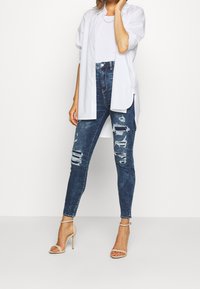 Camisa blanca de gran tamaño, jeans ajustados de mezclilla oscura desgastados y sandalias de tacón nude. Detalles de desgaste notables en los jeans.