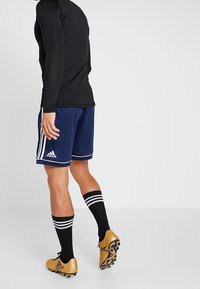 Haut noir à manches longues, short bleu avec des rayures blanches, chaussettes noires montantes jusqu'aux genoux, et chaussures de football dorées avec des accents noirs.