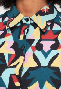 Chemise à manches courtes présentant un motif géométrique audacieux en bleu sarcelle, rouge, jaune et rose, avec un bouton noir et un col classique.