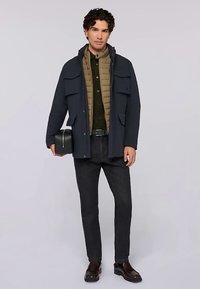 Marinblå jacka med fyra framfickor över en beige quiltad väst, grön skjorta, mörka jeans och bruna läderskor, hålls med en svart väska.