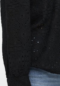 Haut en dentelle noire présentant un motif complexe avec des détails en ajouré. Tissu texturé avec une coupe décontractée, associé à un denim en bas.