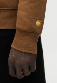 Χέρι με σκουρόχρωμο δέρμα, σφιγμένο χαλαρά, φοράει καφέ ριγέ μανσέτα φούτερ με μικρό κίτρινο κεντημένο λογότυπο κοντά στον καρπό.