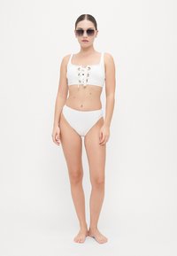 White bikini set featuring a crop top with lace-up front and gold eyelets; high-waisted bottom; smooth fabric; minimalist design.