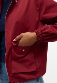 Main placée dans la poche avant à rabat d'une veste rouge avec poignet élastique et fermeture éclair argentée, portée sur un haut côtelé foncé et un jean bleu clair.