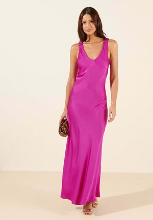 Femme portant une robe de soirée en satin rose vif sans manches, tenant une pochette métallique bronze, debout sur un fond clair uni.