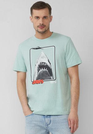 Light blue cotton t-shirt featuring a black-and-white shark graphic with red "JAWS" text and "1975" below, crew neck, short sleeves.