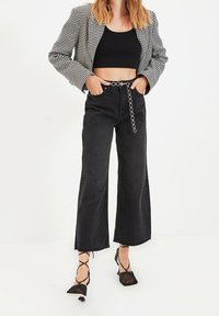 Jean noir à jambes larges avec ourlet brut et détail chaîne à la taille, associé à un top court noir et un blazer à carreaux.