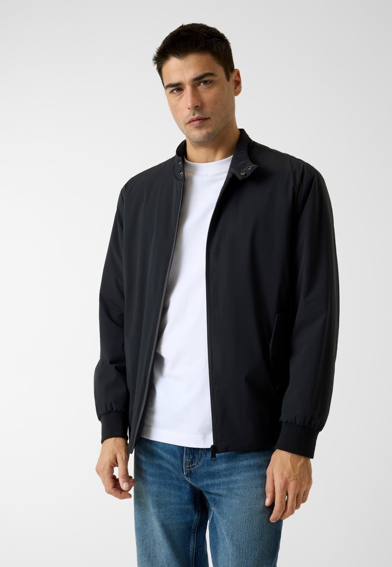 Veste bomber noire avec col montant, fermeture éclair frontale et poignets élastiques. Portée avec un t-shirt blanc et associée à un jean bleu.