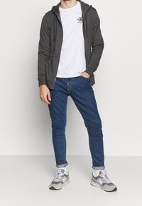 Ein Mann trägt einen grauen Zip-Hoodie, ein weißes T-Shirt mit Grafik, hochgekrempelte blaue Jeans, weiße Socken und graue New Balance Turnschuhe und steht.
