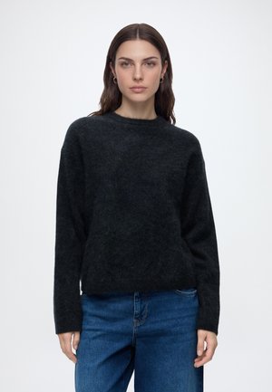 Vrouw met lang bruin haar draagt een zwarte pluizige trui en een blauwe spijkerbroek, staand tegen een effen witte achtergrond.