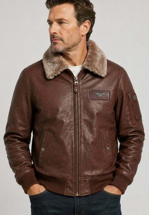 Homme portant une veste aviateur en cuir marron avec col en fourrure, fermeture éclair à l'avant, écusson sur la poitrine et mains dans les poches, regardant vers la gauche.