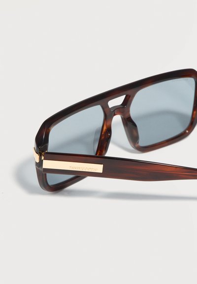 Σκούρα ορθογώνια γυαλιά ηλίου σε σχέδιο χελώνας με μπλε φακούς και χρυσές λεπτομέρειες στους βραχίονες, απεικονισμένα πάνω σε ανοιχτόχρωμο φόντο.