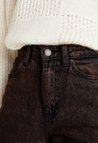 Jeans de denim castanho-escuro com um corte clássico, apresentando um fecho frontal por botão e uma textura subtil. Um suéter tricotado em creme cobre parcialmente os jeans.