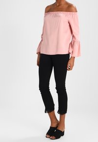 Blusa rosa off-the-shoulder con scollatura arricciata e maniche a campana, abbinata a jeans skinny neri con orlo a frange e mule neri open-toe.