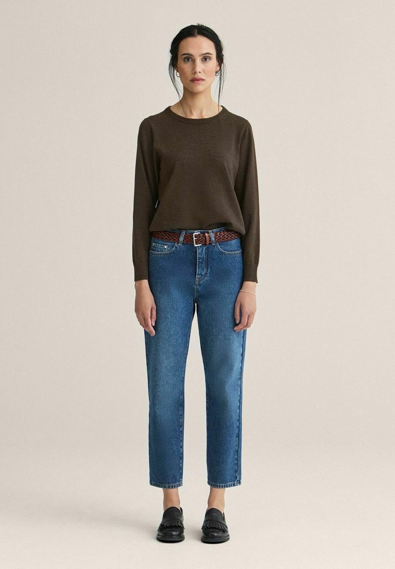 Ruskea pitkähihainen neulepaita, siniset korkeavyötäröiset farkut vaalealla pesulla, punottu ruskea vyö ja mustat loaferit. Yksinkertainen muotoilu, rento istuvuus.