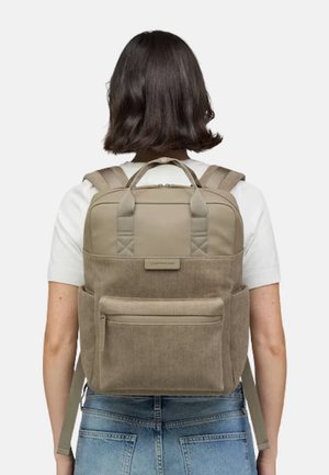 Frau mit beigem Cord-Rucksack mit Frontreißverschlusstasche und oberen Griffen, gekleidet in weißem Hemd und blauer Jeans, von hinten gezeigt.