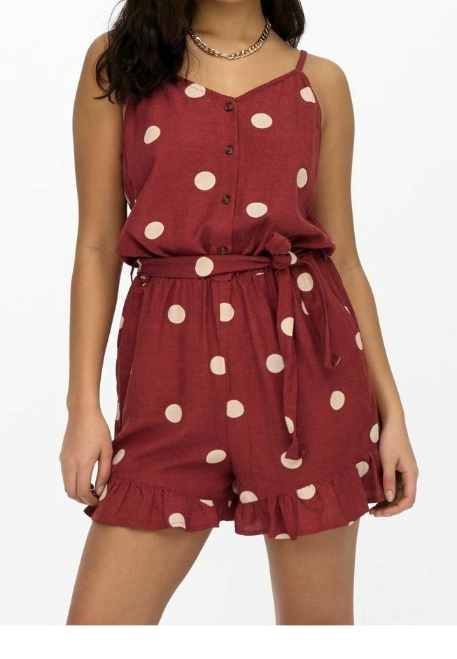 Femme portant une combinaison sans manches bordeaux à pois blancs, avec des boutons à l'avant, une ceinture à la taille et un short à ourlet volanté.