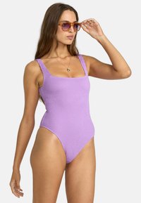 Maillot de bain une pièce violet à côtes avec un décolleté carré et des bretelles larges, mettant en valeur un design de jambe haute. Les accessoires incluent des lunettes de soleil teintées orange.