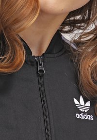 Svart idrotts zip-up jacka tillverkad av slätt tyg, med hög krage, vitt Adidas-logotyp och dragkedja med svart dragflik.