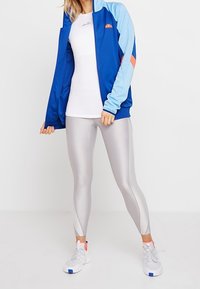 Blå idrottsjacka med ljusblå och korallaccent, vit åtsittande topp, silverleggings och vita sneakers med blå detaljer.