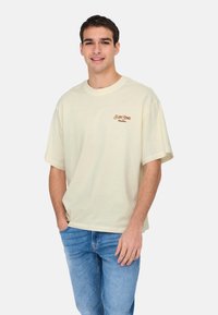 Kræmfarvet bomulds T-shirt med korte ærmer, rund halsudskæring og broderet "Slim Times Panama"-logo i brunt på brystet.